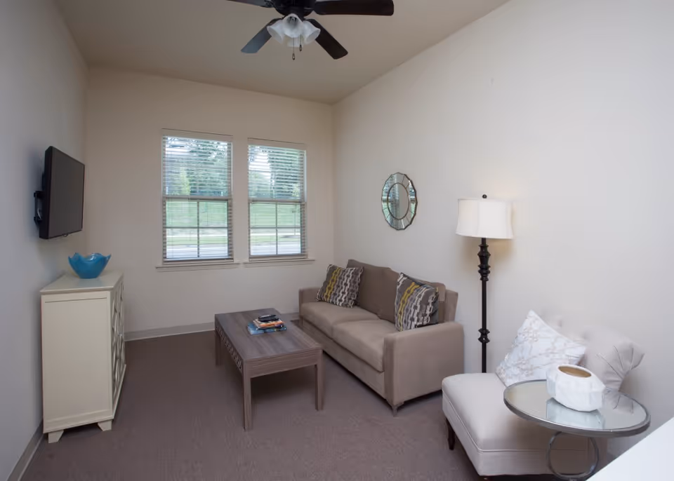 Small furnished living room with a sofa, armchair, coffee table, wall-mounted TV, and a window.