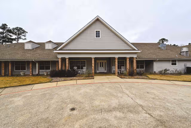 Front exterior view of a single-story building with a peaked roof and a covered entrance supported by brick columns. The building is surrounded by a concrete driveway and some grass areas with a few trees in the background under a cloudy sky.