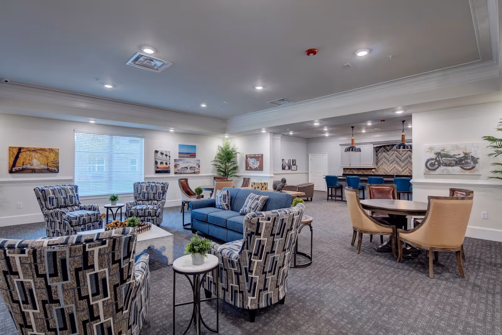 Bright communal lounge with upholstered chairs, a sofa, small tables, a dining table and a bar area.