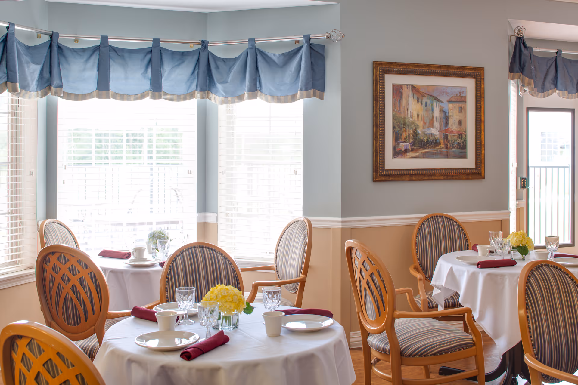 A bright dining room with round tables covered in white tablecloths, set with plates, cups, glasses, and burgundy napkins. Wooden chairs with striped cushions surround the tables. Large windows with blue valances allow natural light to fill the room. A framed painting hangs on the wall.