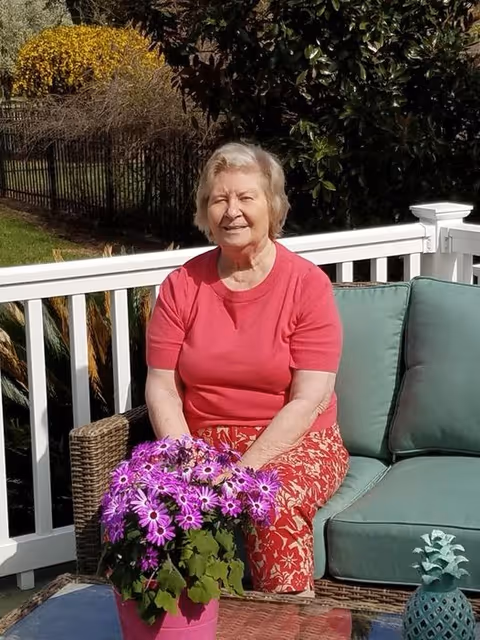 An elderly woman wearing a red shirt and patterned red pants sitting on a wicker outdoor sofa with green cushions on a patio. In front of her is a table with a pink pot of purple flowers. Behind her is a white railing and greenery including bushes and trees.