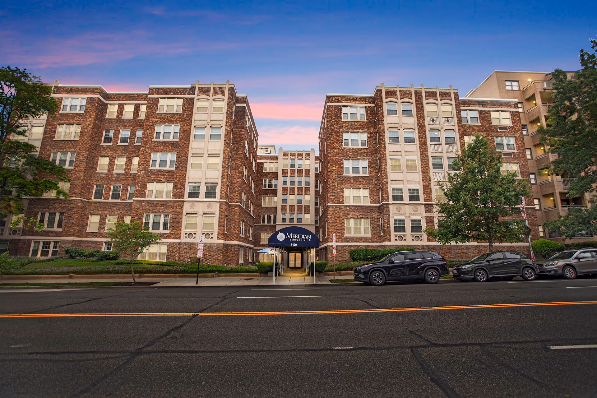 Front exterior view of a large brick senior living building with multiple windows and a central entrance covered by a blue awning that reads 'Meridian Senior Living'. Several cars are parked along the street in front of the building, and trees and shrubs line the sidewalk.