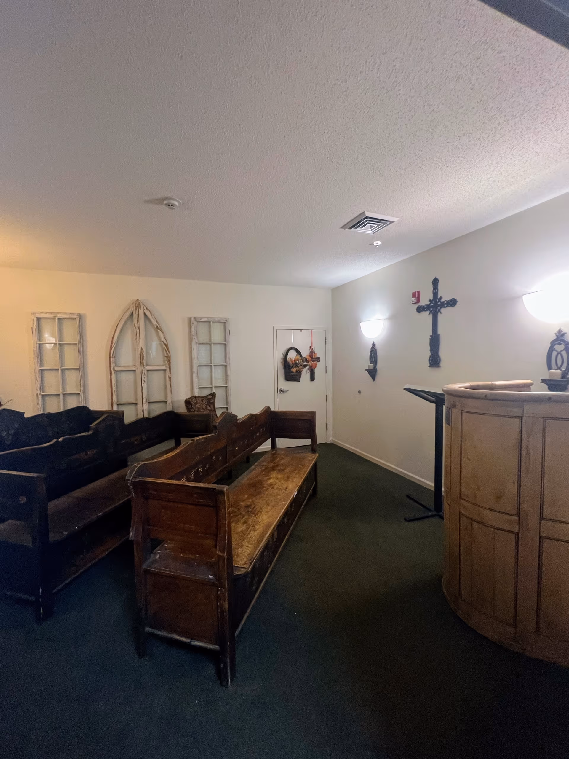Interior room with wooden pew-style benches arranged facing a wooden podium. The walls are decorated with rustic window frames, a cross, and small wall-mounted shelves with decorative items. The room has carpeted flooring and soft wall lighting.