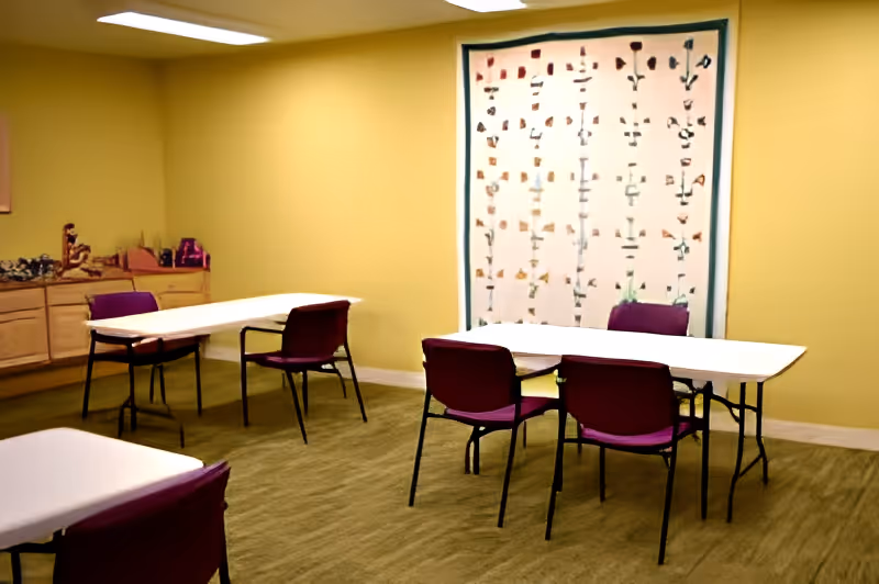 A room with yellow walls and carpeted floor featuring three white rectangular tables and several purple chairs arranged around them. There is a large decorative wall hanging with floral patterns on one wall, and a wooden cabinet with various items on top along another wall. The room is lit by overhead fluorescent lights.