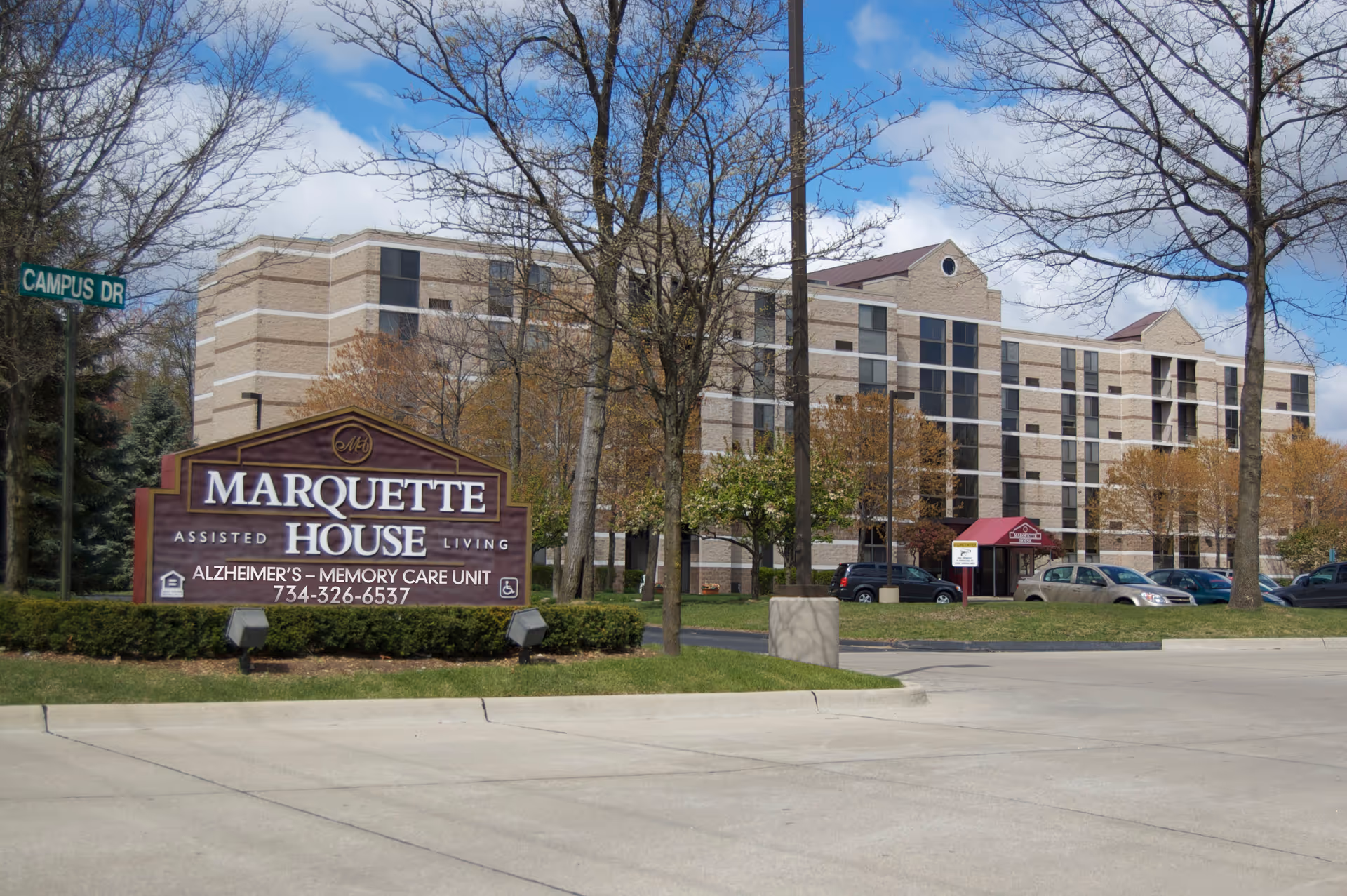 Exterior view of a multi-story assisted living facility named Marquette House, with a large sign in front indicating it offers Alzheimer's memory care. The building is surrounded by trees and parked cars, under a partly cloudy sky.