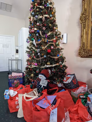 A tall, decorated Christmas tree with colorful ornaments and ribbon stands in a room. Underneath the tree are numerous red and other colored gift bags, some with holiday-themed designs. A snowman decoration holding a sign that says 'LET IT SNOW' is also placed among the gifts. The room has a white door, a metal cart, and a large ornate gold-framed mirror on the wall.