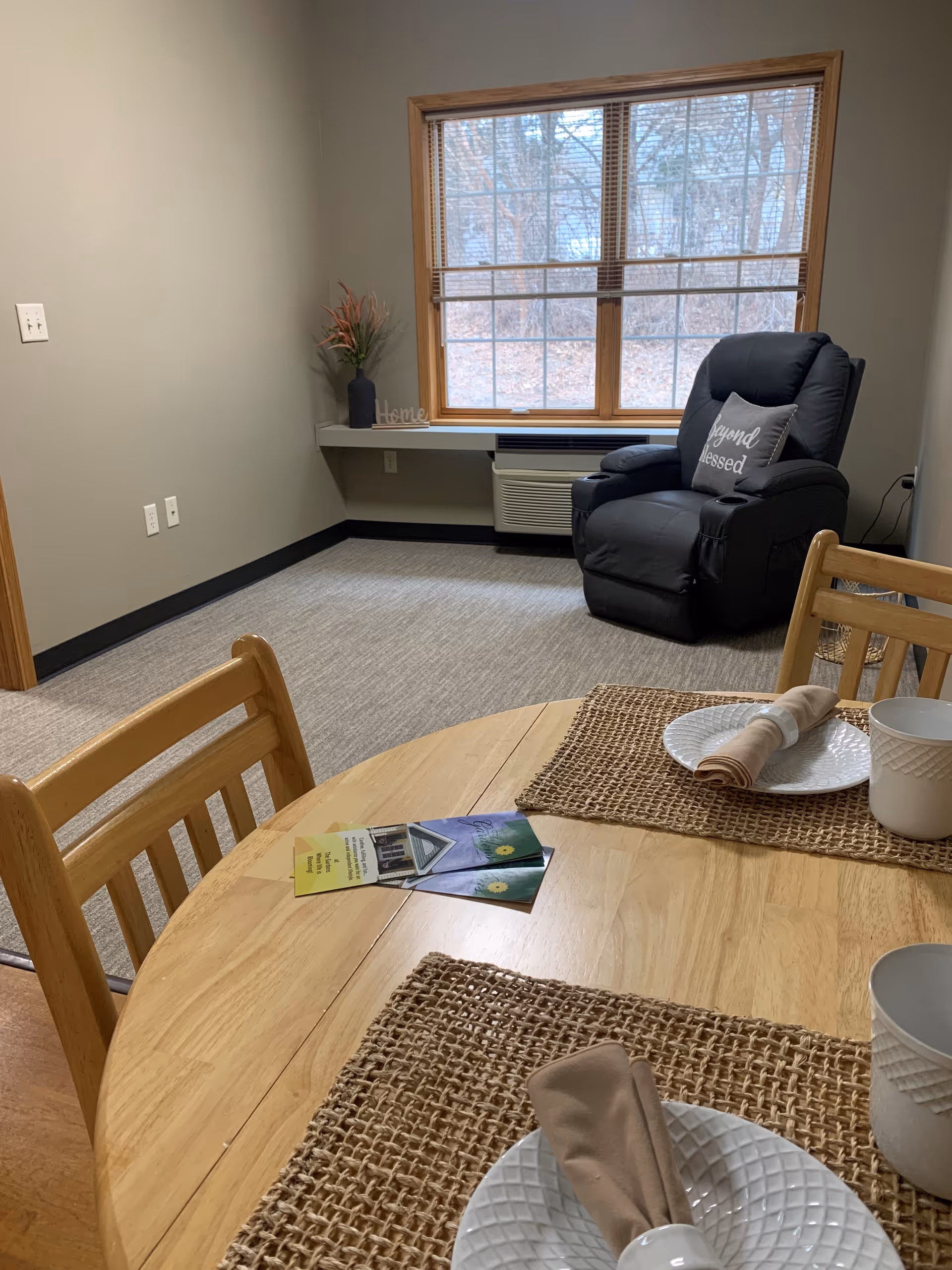 A cozy room with a wooden dining table set for two with white plates, beige napkins, and cups on woven placemats. In the background, there is a large window with blinds, a black recliner chair with a pillow that says 'beyond blessed', a small shelf with a vase of flowers and a decorative 'Home' sign, and a wall-mounted air conditioning unit below the window.