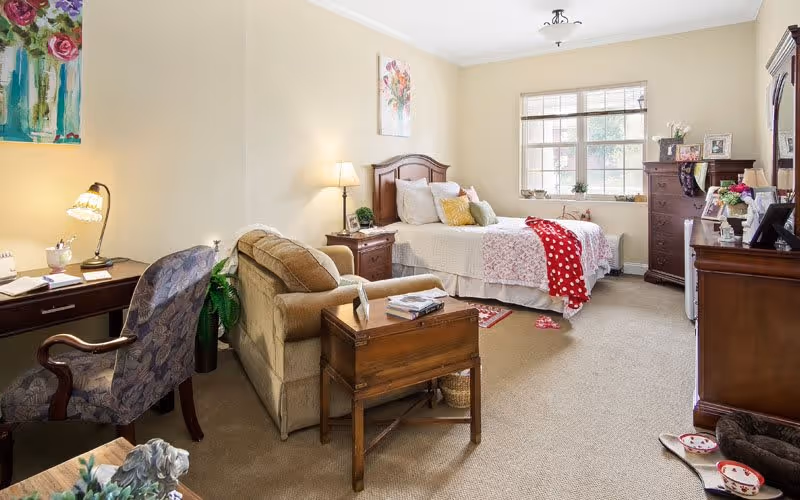A cozy bedroom in a senior living facility featuring a bed with white and floral bedding, a wooden nightstand with a lamp, a comfortable armchair, a wooden desk with a chair and lamp, a large window letting in natural light, and wooden dressers with framed photos and decorations.