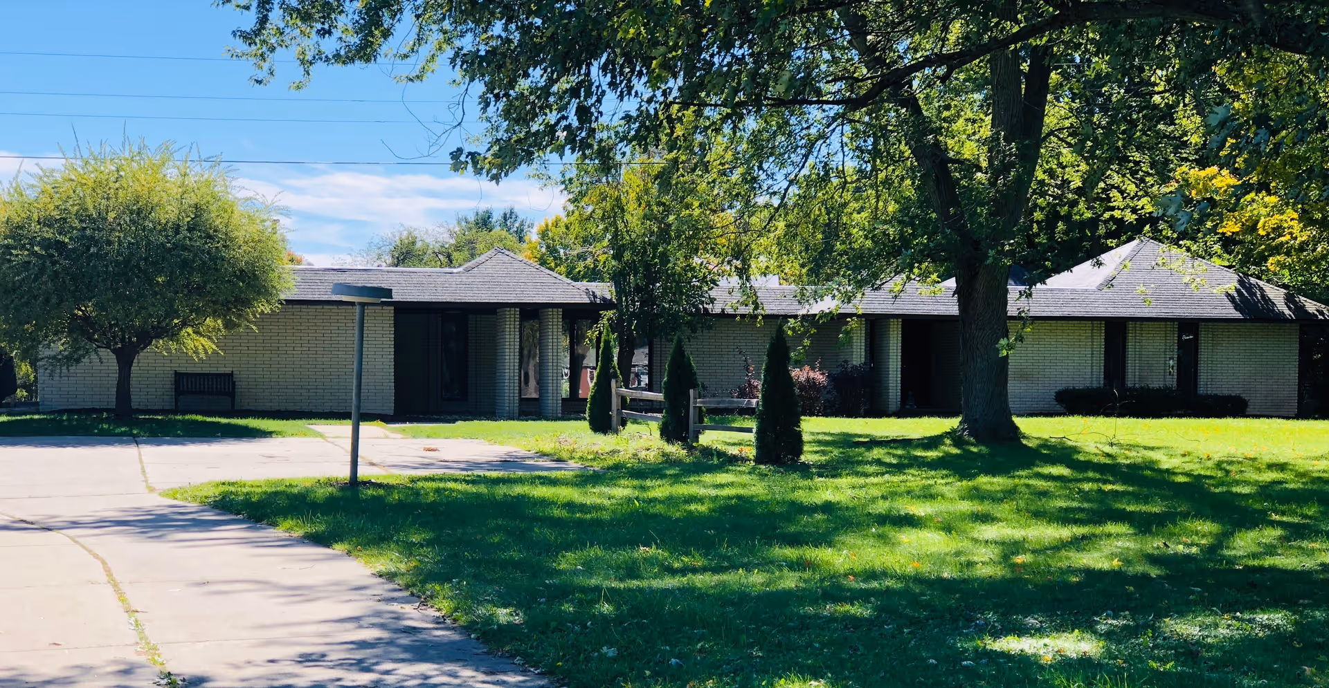 Single-story brick building with a front lawn, trees, and a driveway under a blue sky.