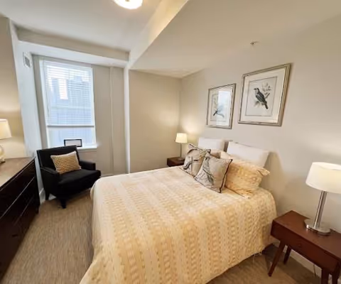 A cozy bedroom with a neatly made bed featuring a beige patterned bedspread and decorative pillows. There are two framed bird illustrations hanging above the bed. On each side of the bed are wooden nightstands with lamps. A black armchair with a patterned cushion is placed near a window with white blinds, and a dresser with a lamp is visible on the left side of the room.