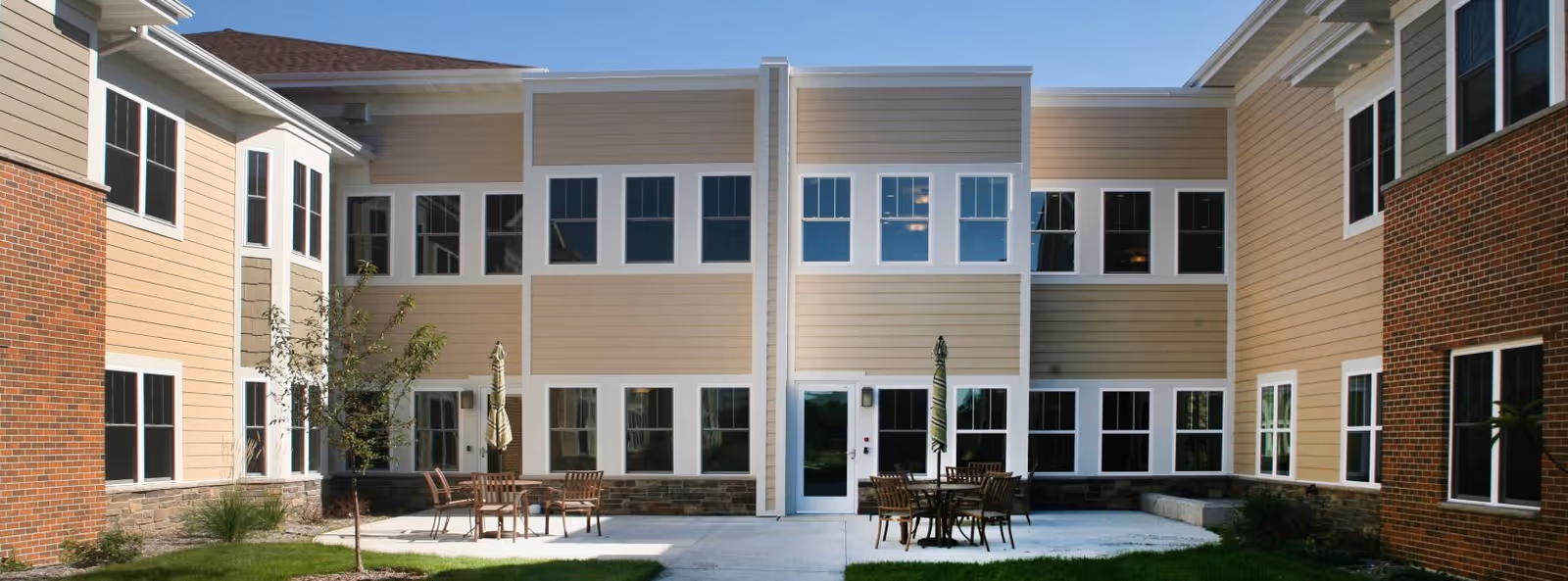 Outdoor courtyard area of a senior living facility with beige and brick exterior walls, multiple windows, patio tables with umbrellas, and small trees and grass surrounding the concrete patio.