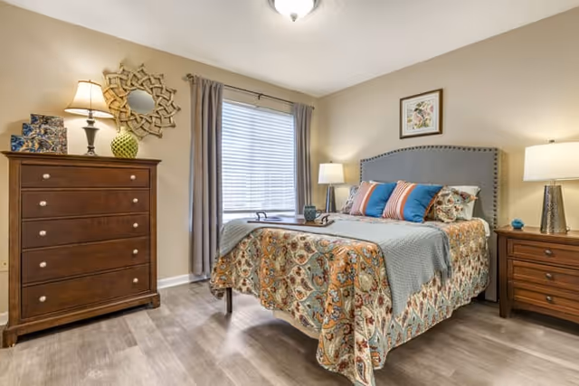 A cozy bedroom with a bed featuring a patterned quilt and multiple pillows. There are two wooden nightstands with lamps on either side of the bed. A wooden dresser with a decorative mirror and a lamp is positioned against the wall. A window with closed blinds and curtains is centered on the back wall, and the room has light-colored walls and wood flooring.