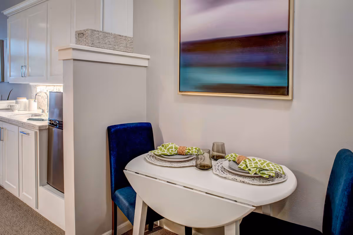 A small dining area with a white drop-leaf table set for two, featuring green patterned napkins and gray plates. Two blue upholstered chairs are placed on either side of the table. In the background, there is a kitchen area with white cabinets, a countertop, and a small refrigerator. A colorful abstract painting hangs on the wall above the table.