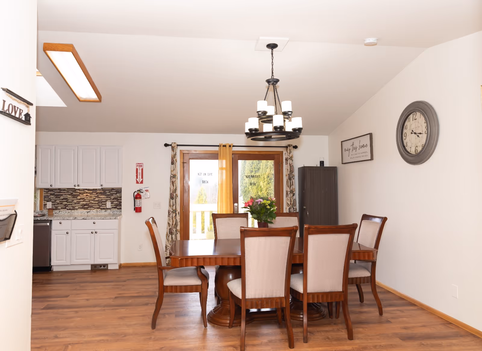 A dining room with a wooden dining table and six upholstered chairs. A chandelier with multiple lights hangs above the table. Behind the table are glass double doors with curtains, leading to an outdoor deck. To the left, part of a kitchen with white cabinets and a tiled backsplash is visible. The room has wooden flooring and white walls, with a large clock and a framed sign on the right wall.