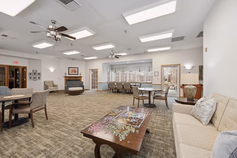 A spacious common area in a senior living facility featuring multiple seating arrangements including armchairs, a sofa, and tables. The room has carpeted floors, ceiling fans, and bright overhead lighting. A fireplace is visible on one wall, and large windows with valances allow natural light into the room. A coffee table in the foreground has a partially completed jigsaw puzzle on it.
