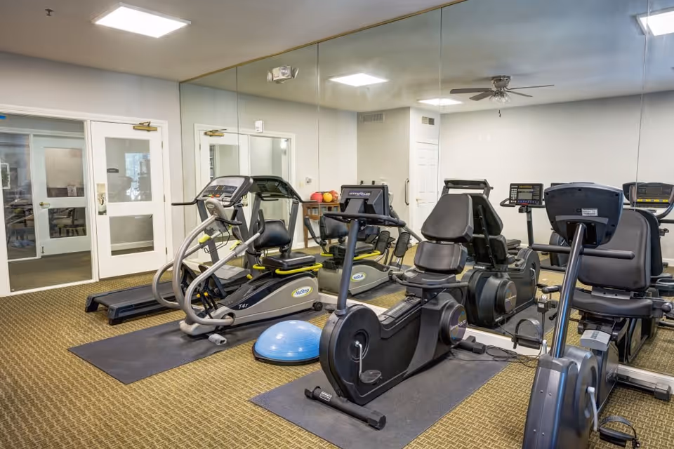Indoor fitness room with exercise equipment including a treadmill, recumbent bikes, and an elliptical machine. The room has a large wall mirror, carpeted floor, and double doors leading to another area.