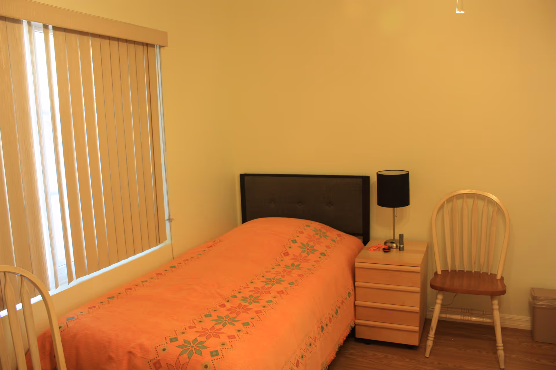 Small bedroom with a single bed, peach bedspread, nightstand, lamp, and chair.