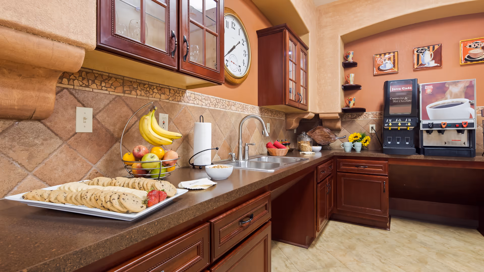 A kitchen area with brown countertops and wooden cabinets. On the counter, there is a plate of assorted cookies with strawberries, a bowl of cereal, a fruit basket with bananas, apples, and oranges, a paper towel holder, and a double sink with a faucet. On the right side, there are two beverage dispensers labeled Java Café for cappuccino, hot chocolate, and coffee. The walls are decorated with coffee-themed artwork and small shelves holding colorful mugs. A large clock is mounted on the wall above the sink.