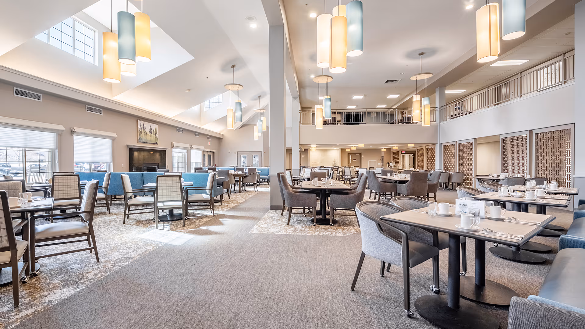 A spacious and well-lit dining room with multiple tables and chairs arranged neatly. The room features high ceilings with large windows allowing natural light to fill the space. Modern pendant lights hang from the ceiling, and the tables are set with cups, plates, and utensils. There is a fireplace along one wall and a second-floor balcony overlooking the dining area.