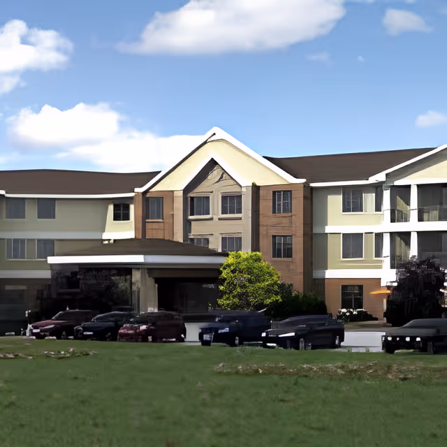 Exterior view of a multi-story senior living facility building with a covered entrance, several windows, and a grassy area in the foreground under a partly cloudy sky.