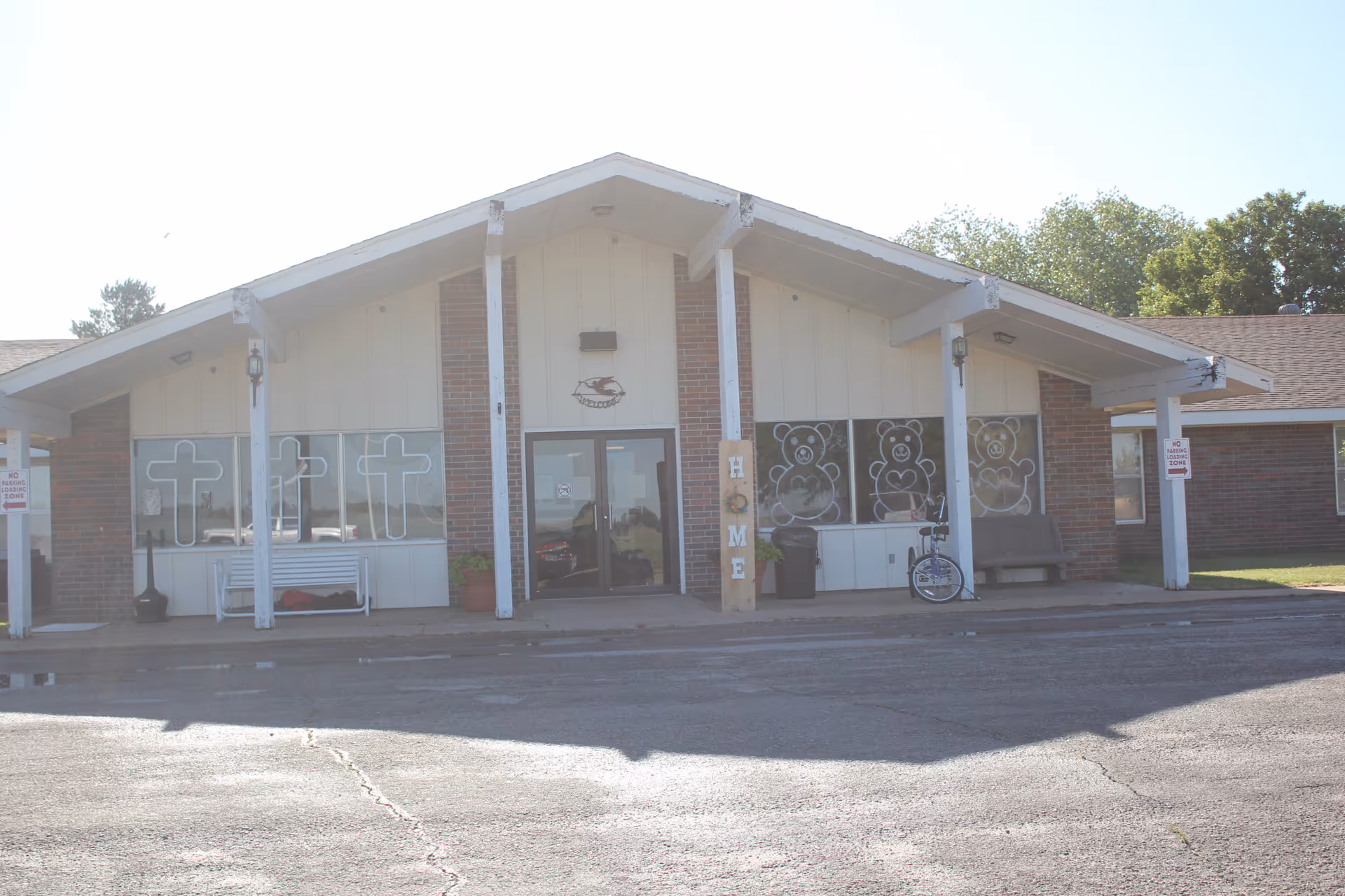 Front exterior view of Protection Valley Manor building with a peaked roof, brick and white panel walls, large windows decorated with cross and teddy bear designs, a bench, a bicycle, and signs indicating no loitering zones.