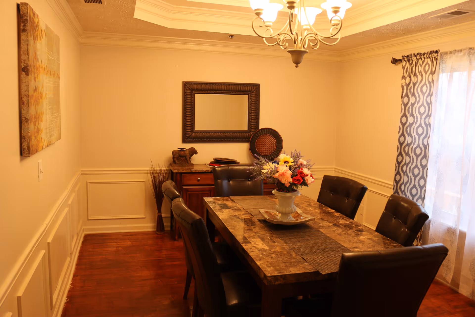 A warmly lit dining room with a rectangular marble-top table surrounded by six dark leather chairs. A vase with colorful flowers is placed in the center of the table. On the far wall, there is a wooden sideboard with decorative items including a small sculpture and a woven plate, above which hangs a framed mirror. A chandelier with five lights hangs from the ceiling. The room has wooden flooring, cream-colored walls with wainscoting, a patterned curtain on the window, and a piece of artwork on the left wall.