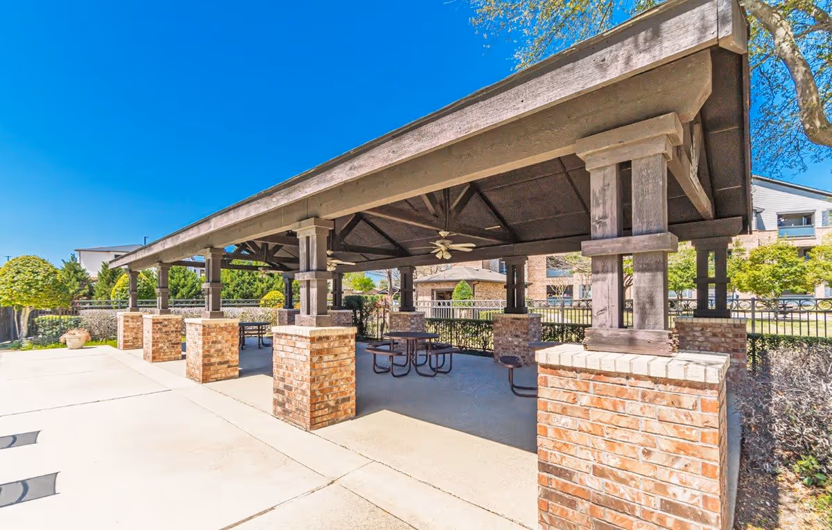 Outdoor covered pavilion with brick pillars and wooden beams, featuring ceiling fans and picnic tables underneath. Surrounding the pavilion are trees, shrubs, and a clear blue sky.