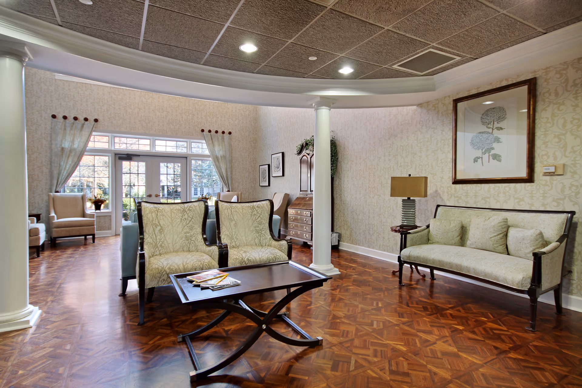 A well-lit living room area with patterned wallpaper, wooden parquet flooring, and a curved ceiling with recessed lighting. The room features two upholstered armchairs with a small dark wooden coffee table in front, a matching upholstered sofa with cushions, a side table with a lamp, framed botanical artwork on the wall, and large windows with sheer curtains letting in natural light.