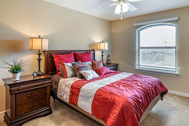 A bedroom with a bed featuring a red, white, and beige comforter and multiple pillows. There are two wooden nightstands on either side of the bed, each with a lamp. A window with blinds is on the right wall, and the room has beige walls and carpeted floor.
