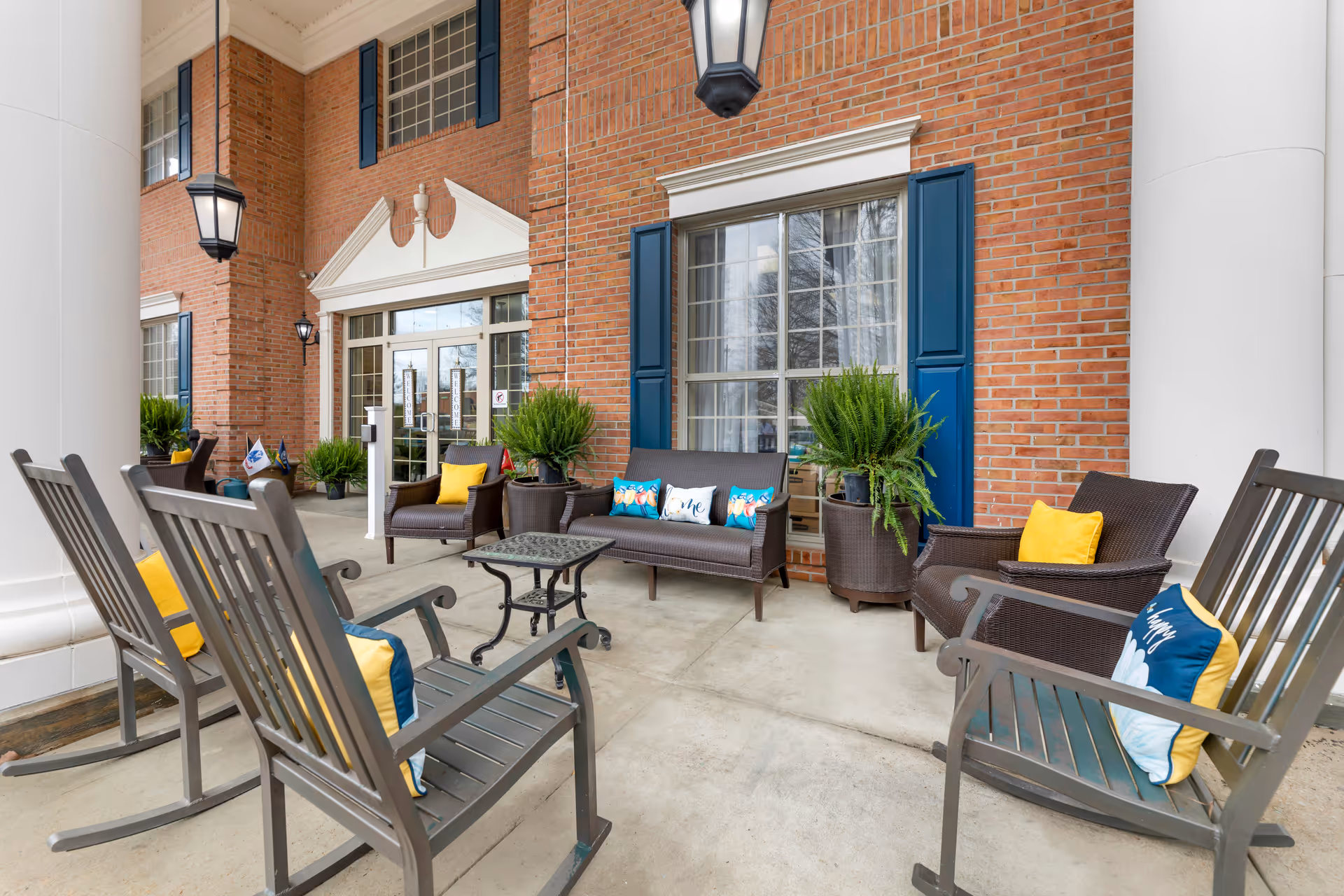 Front entrance porch of a brick building with rocking chairs, wicker seating, colorful cushions and potted plants.