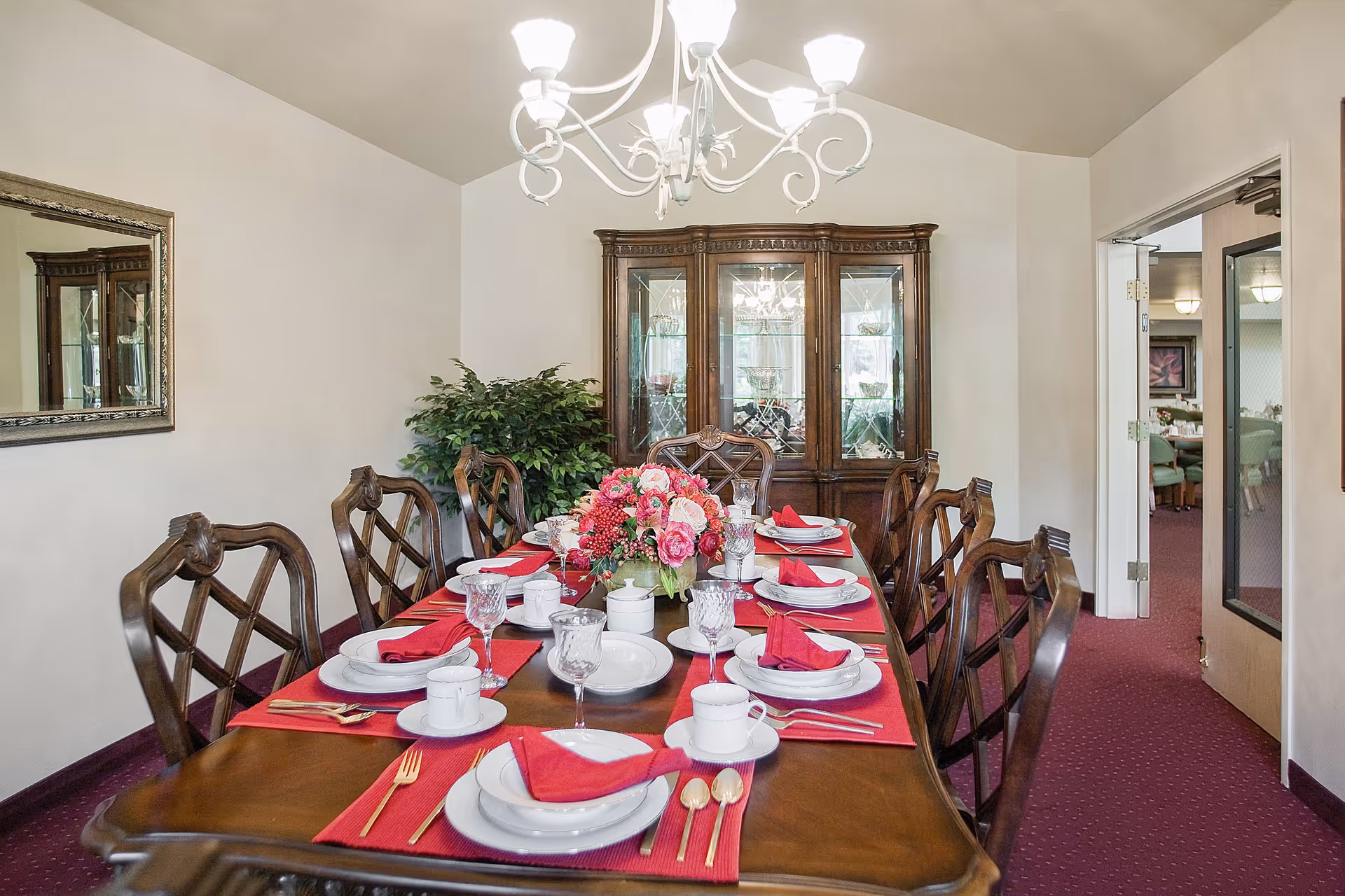 A formal dining room with a long wooden table set for eight people. The table is adorned with red placemats, white plates, red napkins, crystal glasses, and gold-colored cutlery. A floral centerpiece with pink and red flowers is in the middle of the table. Behind the table is a wooden china cabinet with glass doors, and a large mirror hangs on the left wall. The room has white walls, a red carpet, and a white chandelier hanging from the ceiling. An open doorway leads to another room with similar decor.