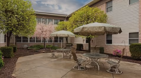 Outdoor courtyard area with round glass tables and chairs under large umbrellas, surrounded by trees and shrubs, adjacent to a two-story building with windows.