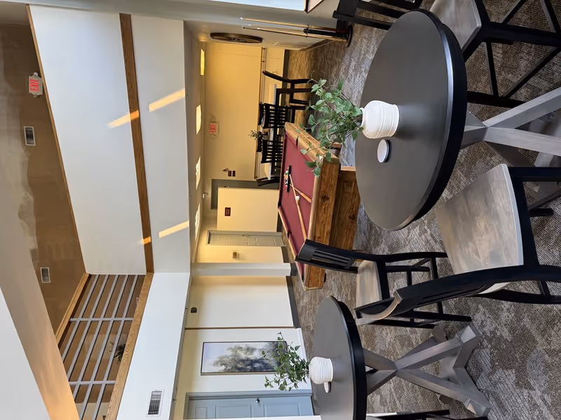 Interior view of a common area in a senior living facility featuring a pool table with cues and balls, round tables with chairs, and potted plants on the tables. The space has a high ceiling with a balcony railing above, neutral-colored walls, and carpeted flooring.