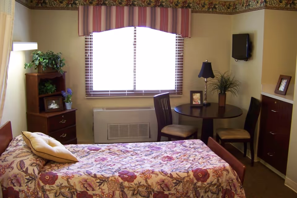 A cozy bedroom in a senior living facility with a single bed covered in a floral quilt and a beige pillow. There is a wooden nightstand with drawers and decorative plants and framed photos on top. A round table with two chairs is positioned near a window with striped valance curtains. A small flat-screen TV is mounted on the wall above a cabinet, and a table lamp and plant are on the round table.