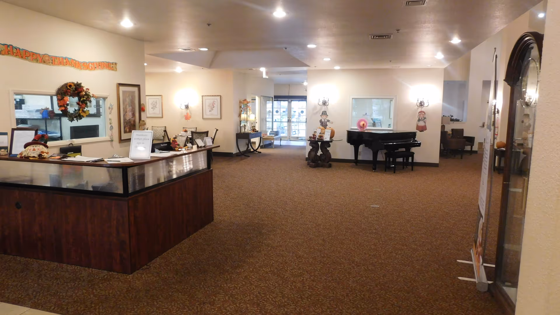 Interior view of a senior living facility lobby area with a reception desk on the left decorated for Thanksgiving, a grand piano in the center back, and various framed pictures and decorations on the walls. The space is well-lit with ceiling lights and has carpeted flooring.