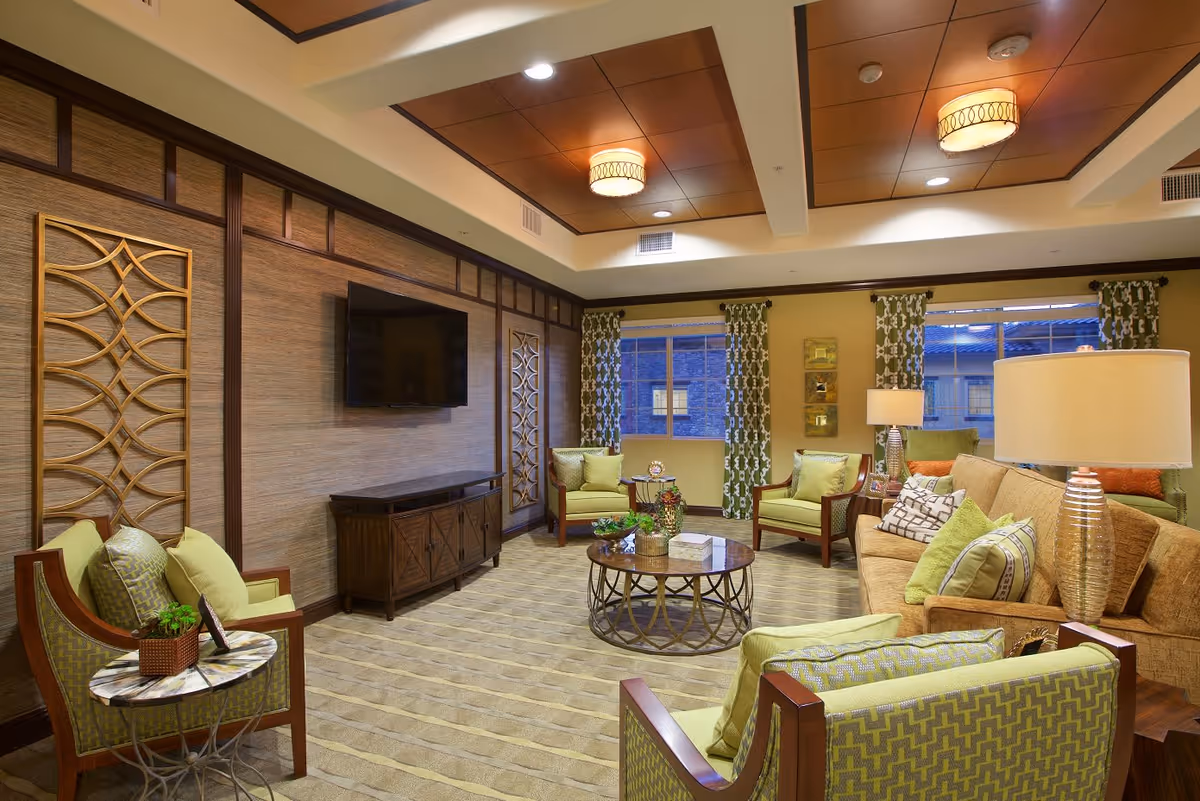 A well-lit living room with green and beige upholstered chairs and a beige sofa adorned with multiple cushions. The room features a round coffee table with decorative items, a wooden TV stand with a flat-screen TV mounted on the wall, patterned curtains on the windows, and warm ceiling lights.