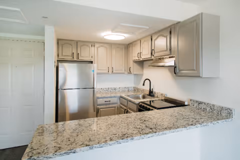 Modern kitchen with granite countertops, beige cabinets, stainless steel refrigerator, electric stove with oven, and a black faucet over the sink. The kitchen has a clean and bright appearance with overhead lighting.