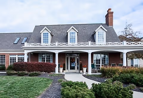 Front exterior view of a brick building with a gray shingled roof, three dormer windows, a chimney, and a covered entrance with white columns. There are shrubs and greenery in the landscaped area in front of the building.