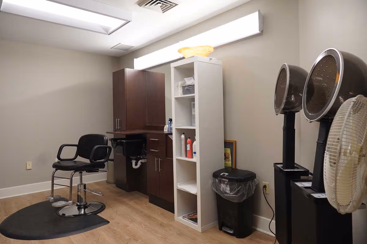Interior view of a salon area in a senior living facility featuring a black salon chair on a black mat, a wooden cabinet with a sink, a white shelving unit with hair care products, two black hair dryer chairs, and a standing fan against beige walls and wooden flooring.