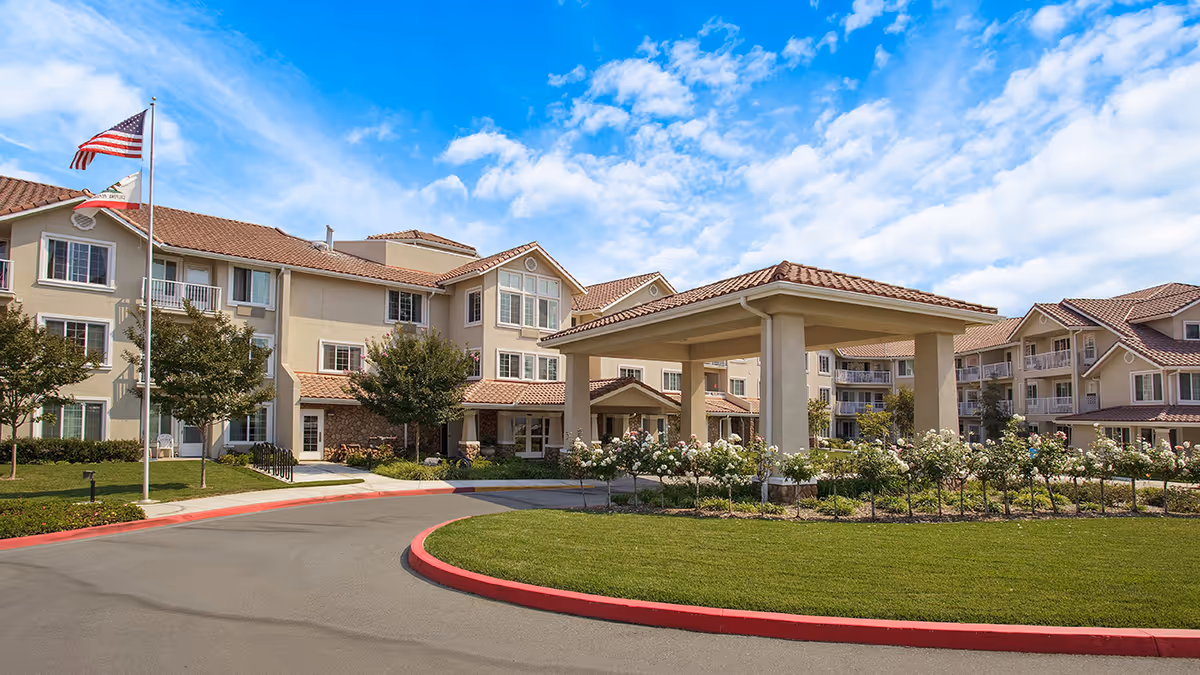 Front exterior of a senior living building with a covered entrance (porte-cochère), landscaped circular drive, and a flagpole under a blue sky.