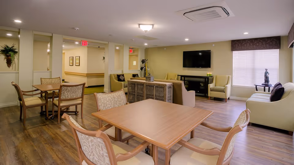 A well-lit common area in an assisted living facility featuring wooden tables with cushioned chairs, a seating area with sofas and armchairs, a flat-screen TV mounted on the wall, and a window with blinds and a decorative valance. The room has wooden flooring and neutral-colored walls with some decorative elements.
