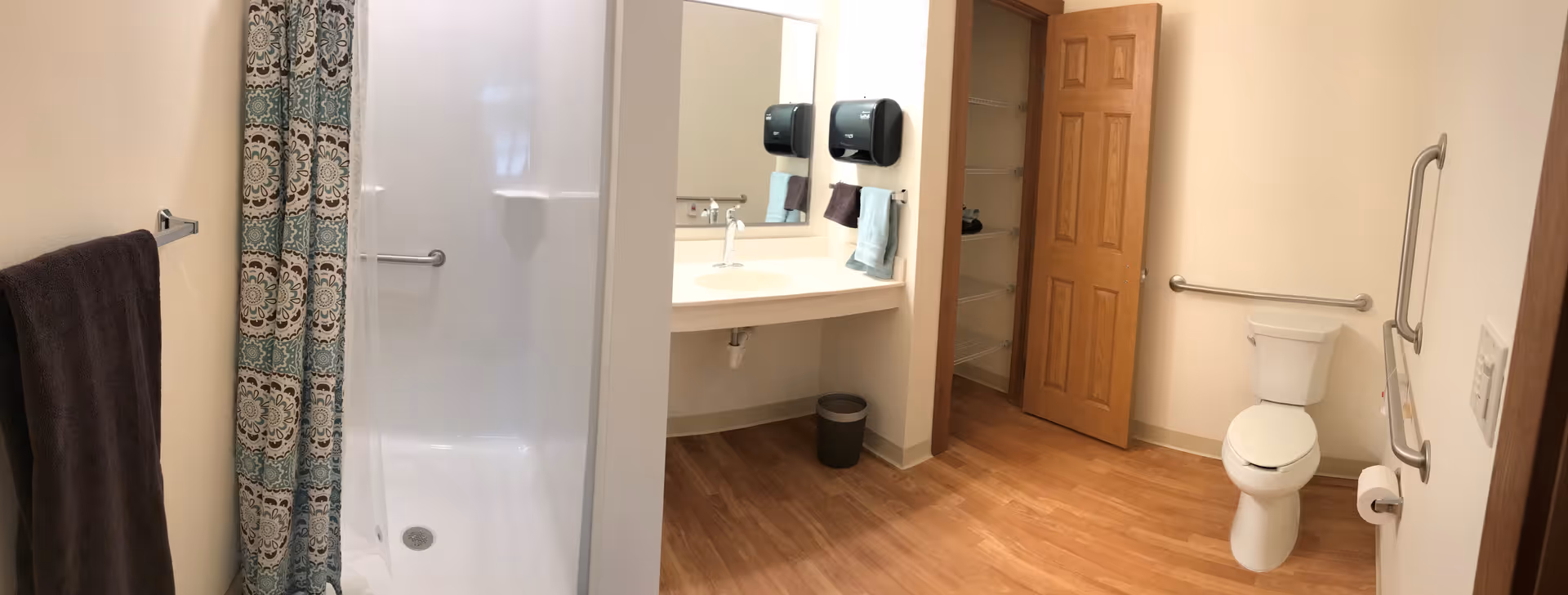 A spacious bathroom with a walk-in shower featuring a patterned shower curtain, a sink with a large mirror above it, two towel holders with towels, a trash bin underneath the sink, a wooden door leading to a closet or storage area, and a toilet with grab bars on the walls for accessibility.