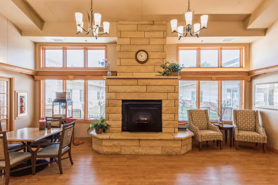 A cozy senior living common area featuring a large stone fireplace with a clock above it, two patterned armchairs with a small round table between them, a dining table with chairs, large windows with wooden blinds letting in natural light, and two ceiling light fixtures.