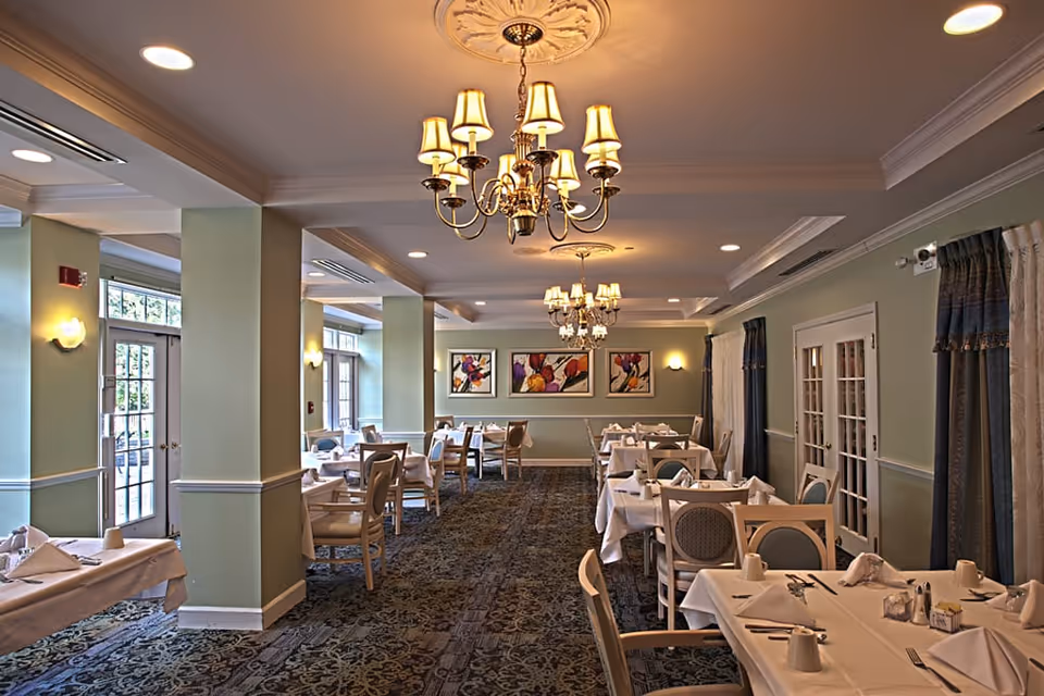 A well-lit dining room with multiple tables covered in white tablecloths, each set with napkins, cups, and silverware. The room features light green walls, decorative chandeliers hanging from the ceiling, patterned carpet, and framed colorful artwork on the far wall. Large windows and glass doors allow natural light to enter the space.