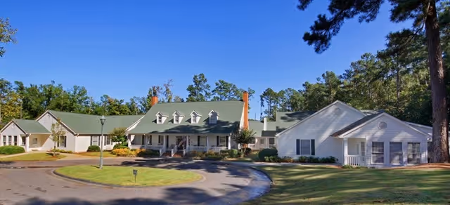 Front exterior view of a large, single-story building with a green roof and white siding, surrounded by trees and a circular driveway under a clear blue sky.