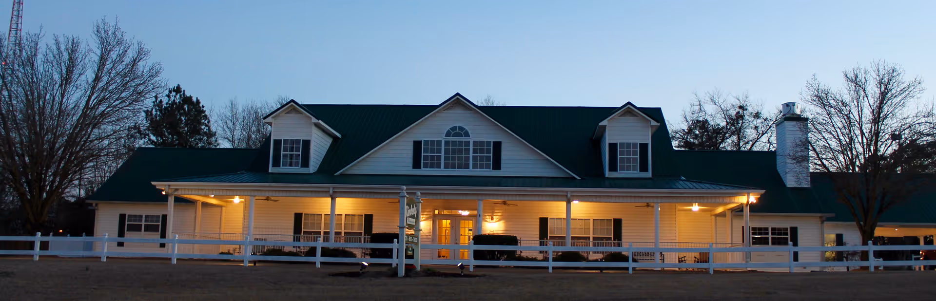 A long care home building with a green roof, white fence, and lit porch.
