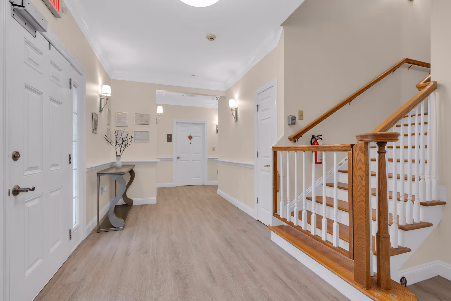 Interior hallway of Serenity Gardens at Brick featuring light beige walls, wood flooring, white doors, a wooden staircase with white balusters, wall-mounted lights, and a small decorative table with a vase and branches.