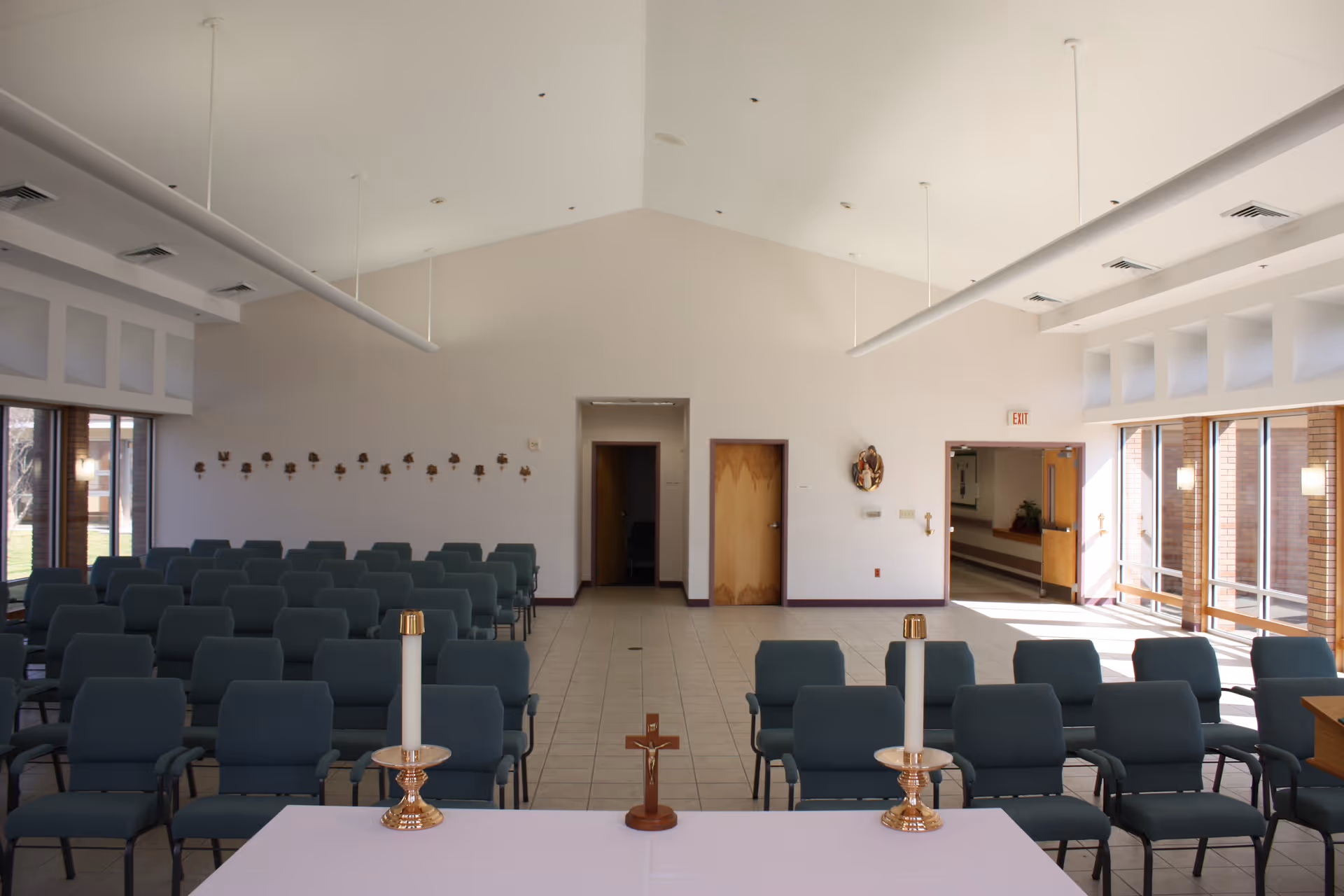 Interior view of a chapel or small worship room with rows of blue chairs facing a white altar table. On the altar are two tall white candles in gold holders and a small wooden crucifix. The room has a high ceiling with hanging lights, large windows on the right side letting in natural light, and two wooden doors at the far end.