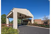 Covered drop-off entrance with a porte-cochère, driveway, and surrounding landscaping in front of a single-story building under a clear blue sky.