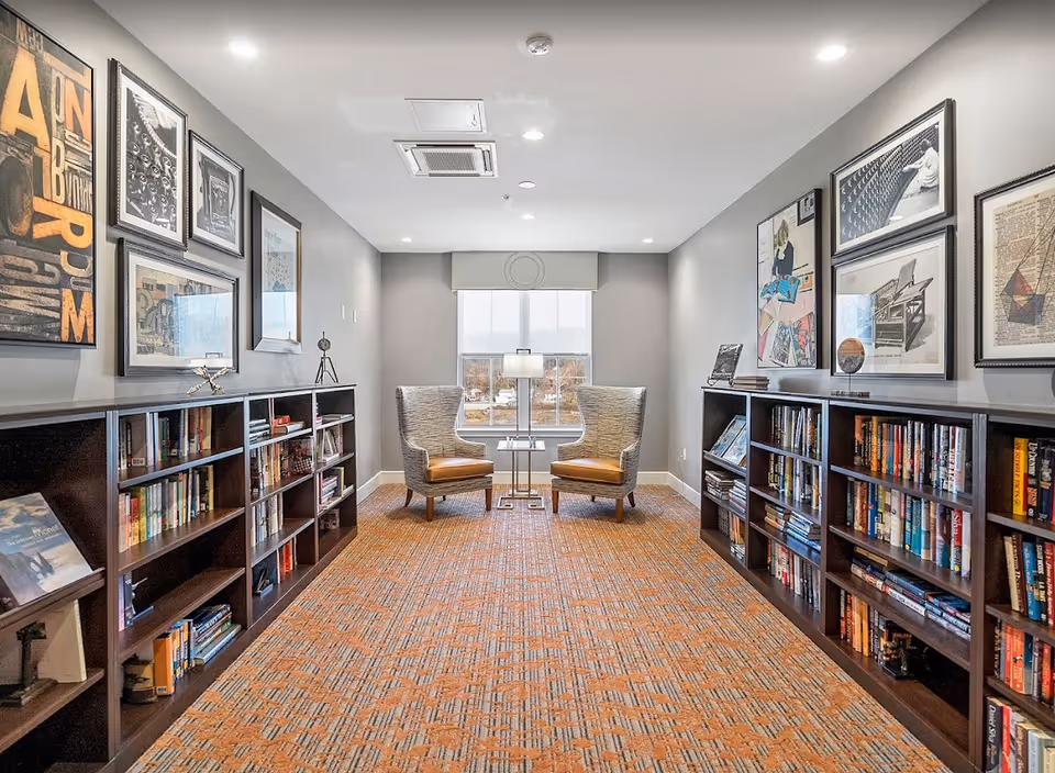 A cozy reading nook in a retirement living facility featuring two cushioned armchairs with wooden legs positioned around a small glass-top table with a lamp. The room has bookshelves filled with books on both sides, framed artwork on the walls, and a large window at the end letting in natural light. The carpet has a patterned design in warm tones.